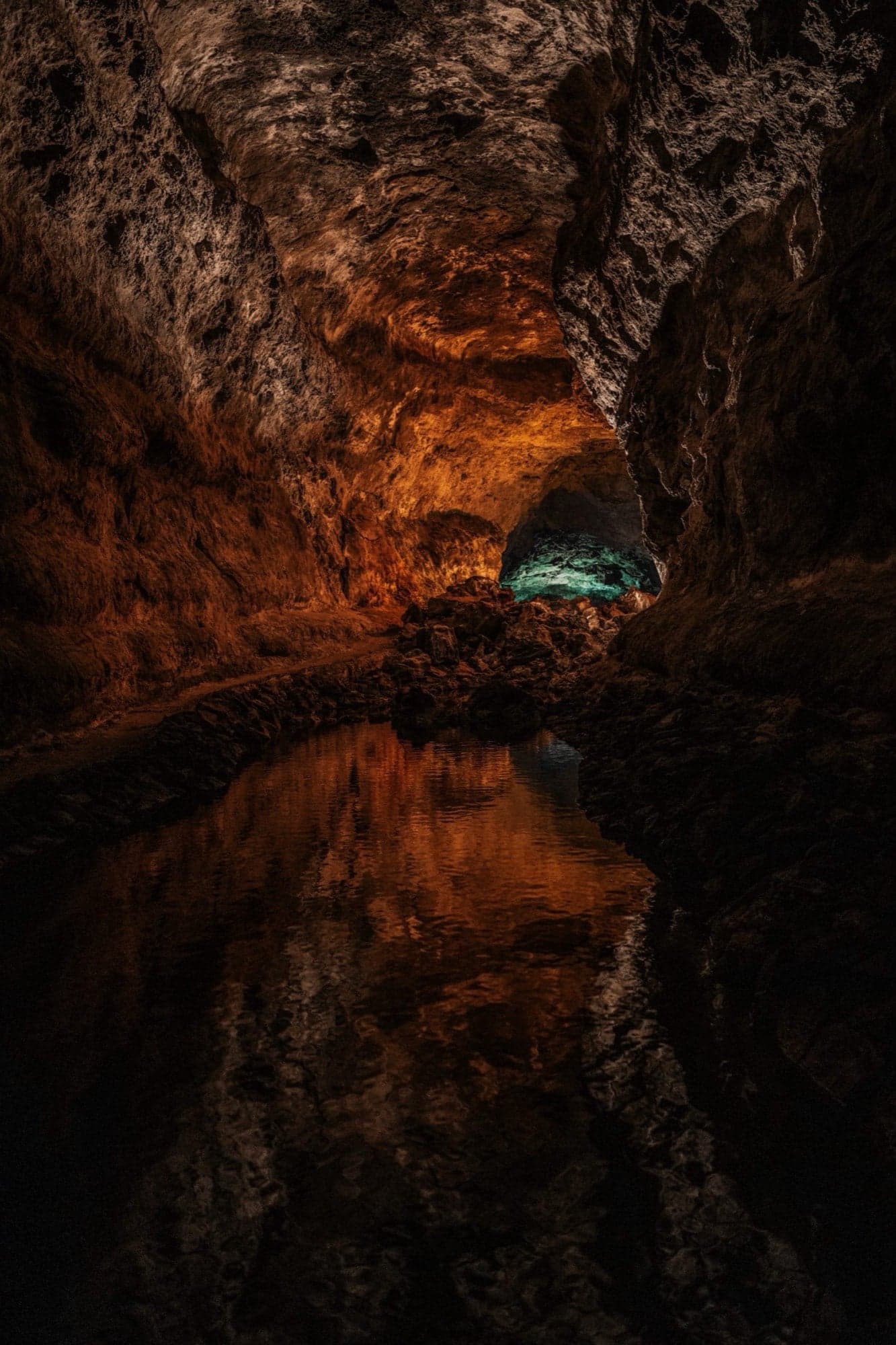 Grottengang met warm oranje verlichting op ruwe lavawanden, turquoise daglicht in de verte en reflectie in stilstaand water