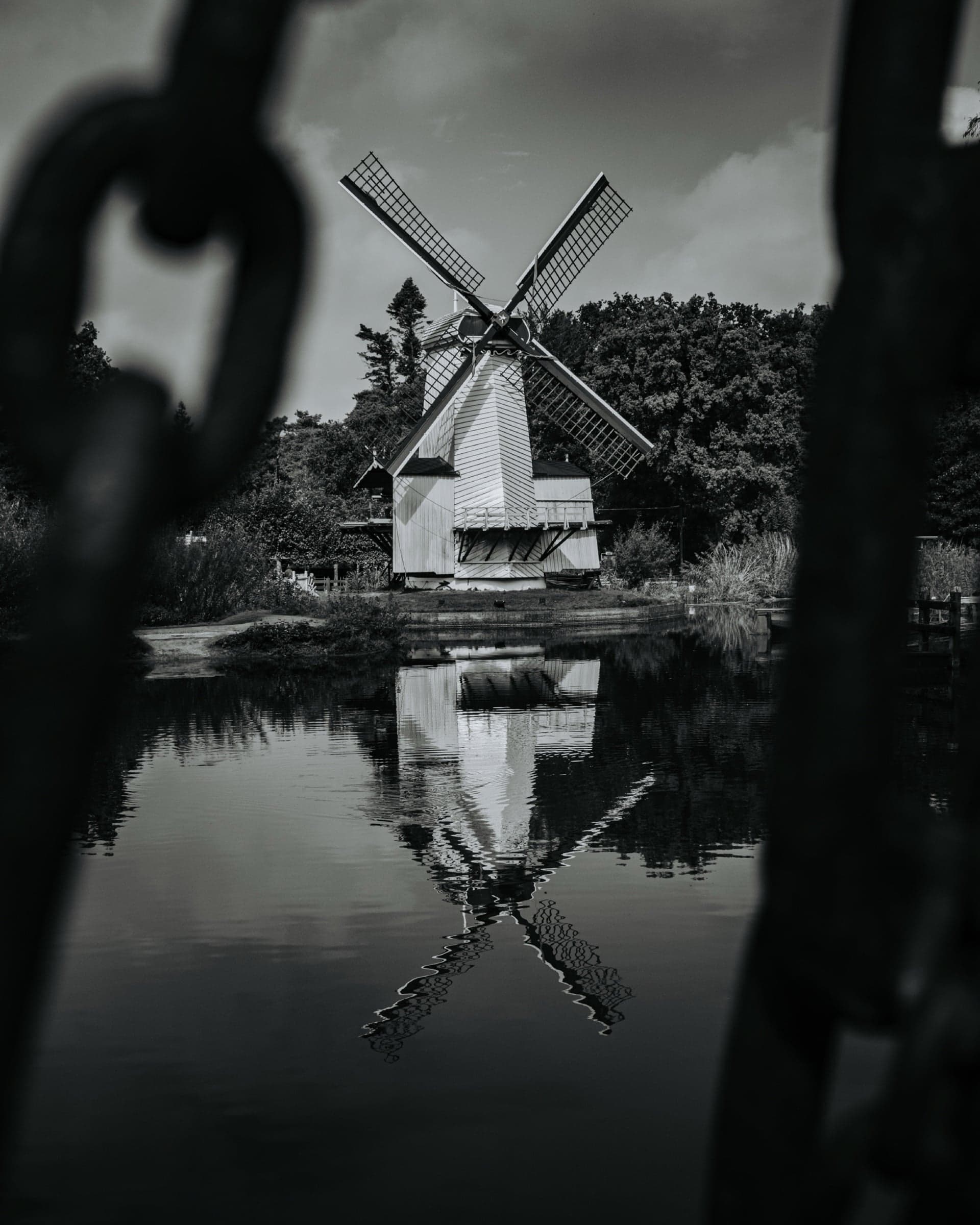 Witte Nederlandse molen in zwart-wit, symmetrisch gespiegeld in stilstaand water, gefotografeerd door een onscherp ijzeren ketting-frame op de voorgrond