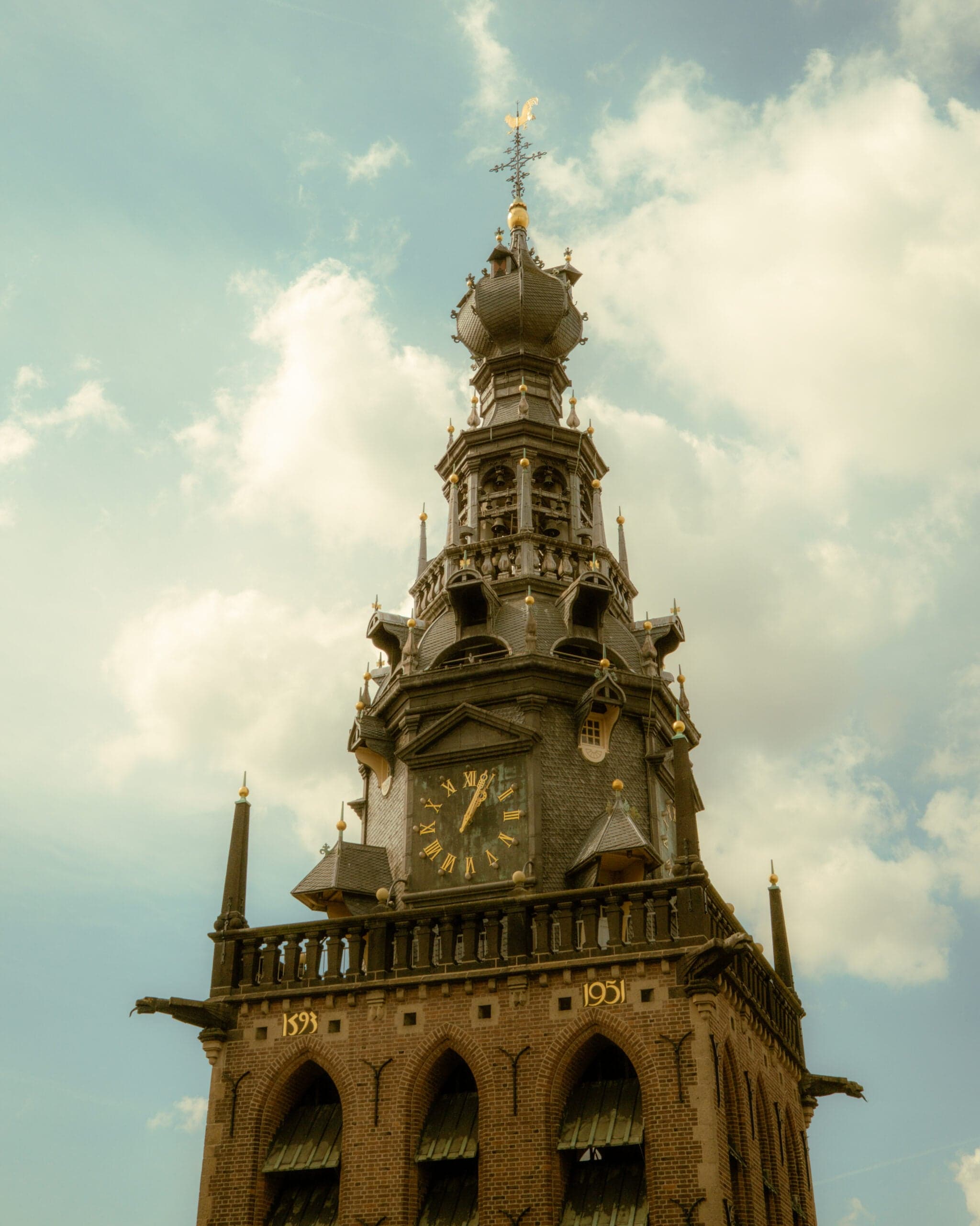 Gotische torenspits van de Stevenskerk in Nijmegen met vergulde windvaan en de jaartallen 1593 en 1931 op de gevel, tegen een lucht met stapelwolken