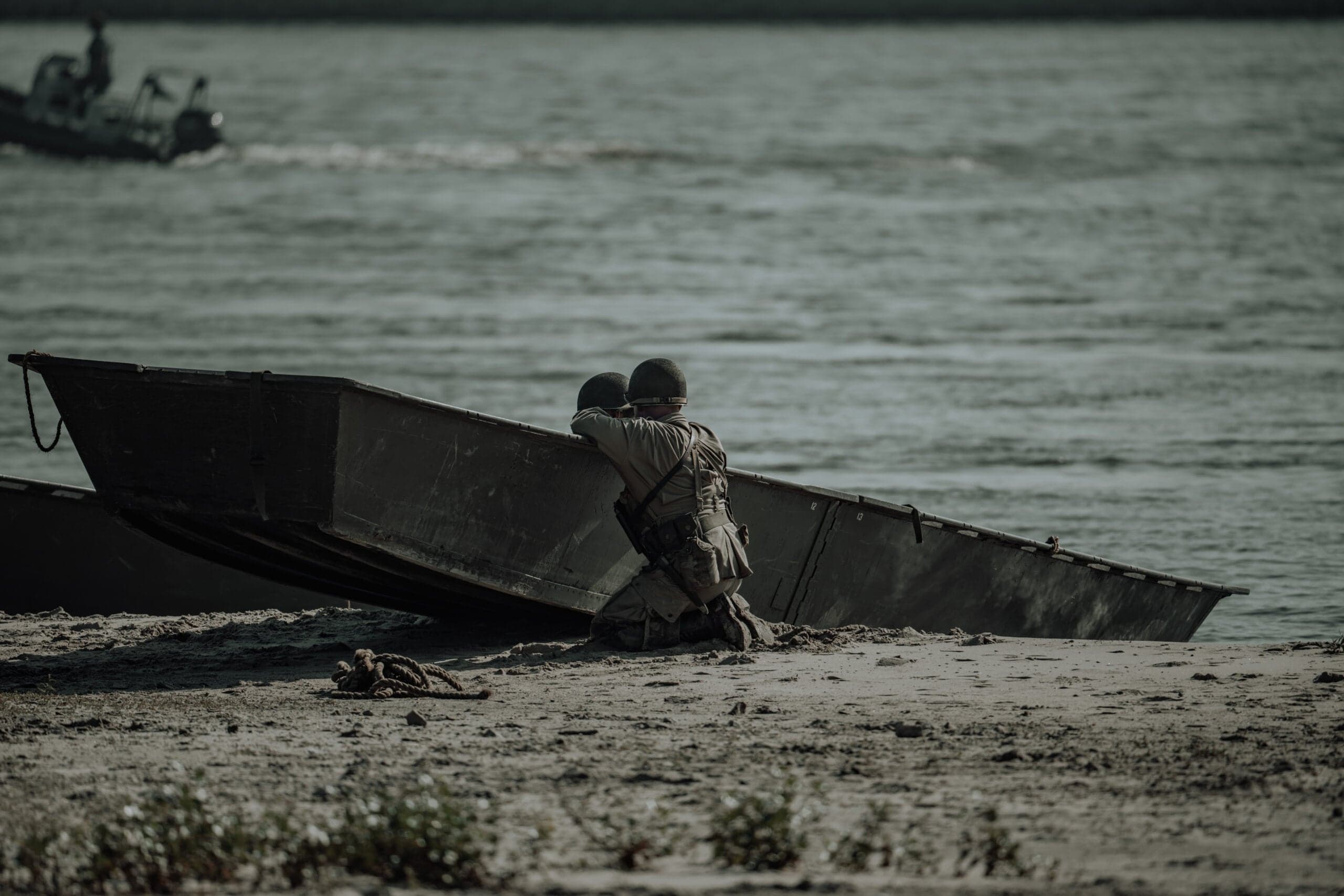 Twee soldaten in WO2-reenactment-uniform gehurkt achter een gestrande stormboot op een zandige rivieroever, nog een vaartuig scheurend over het water op de achtergrond