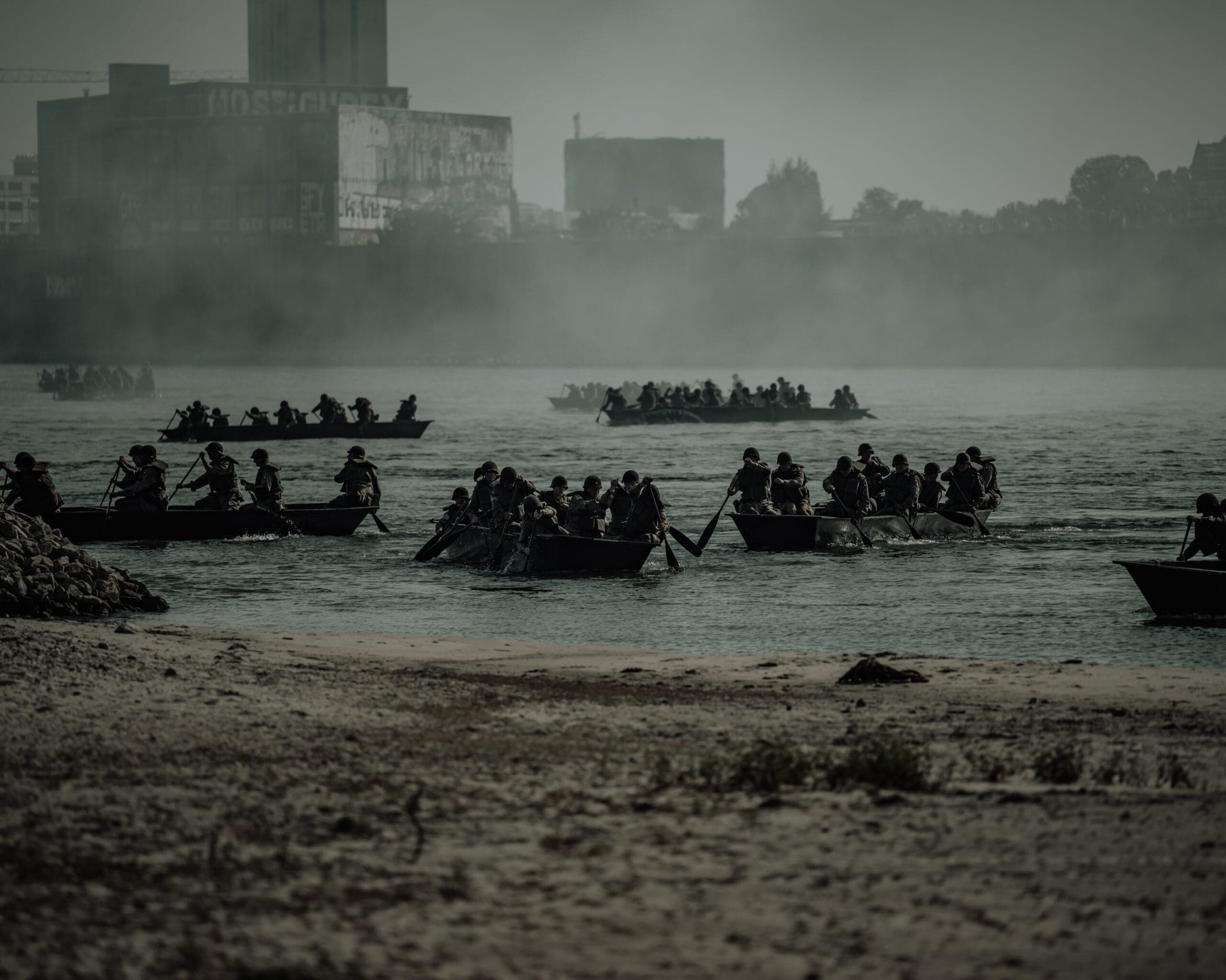 Flottielje assaultboten vol soldaten steekt bij dageraad over een rivier, silhouet van fabriek en stad door de ochtendmist
