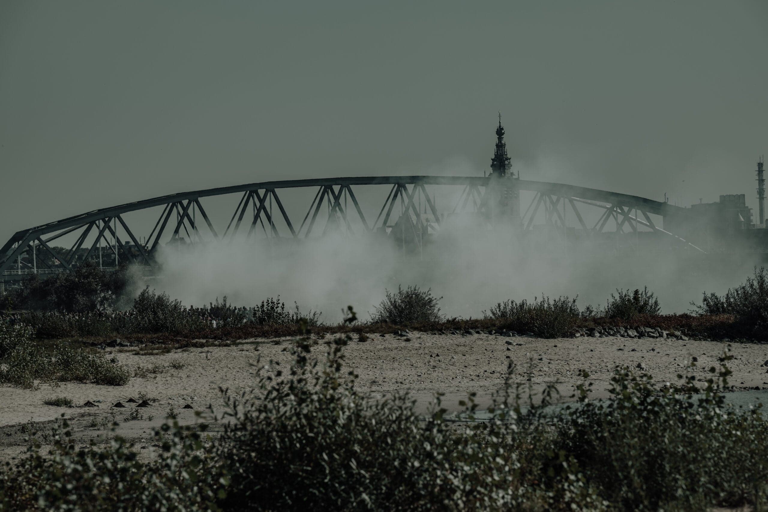 Boogbrug van Nijmegen half verhuld in rookslierten, wilde grassen op de voorgrond onder een melkwitte lucht