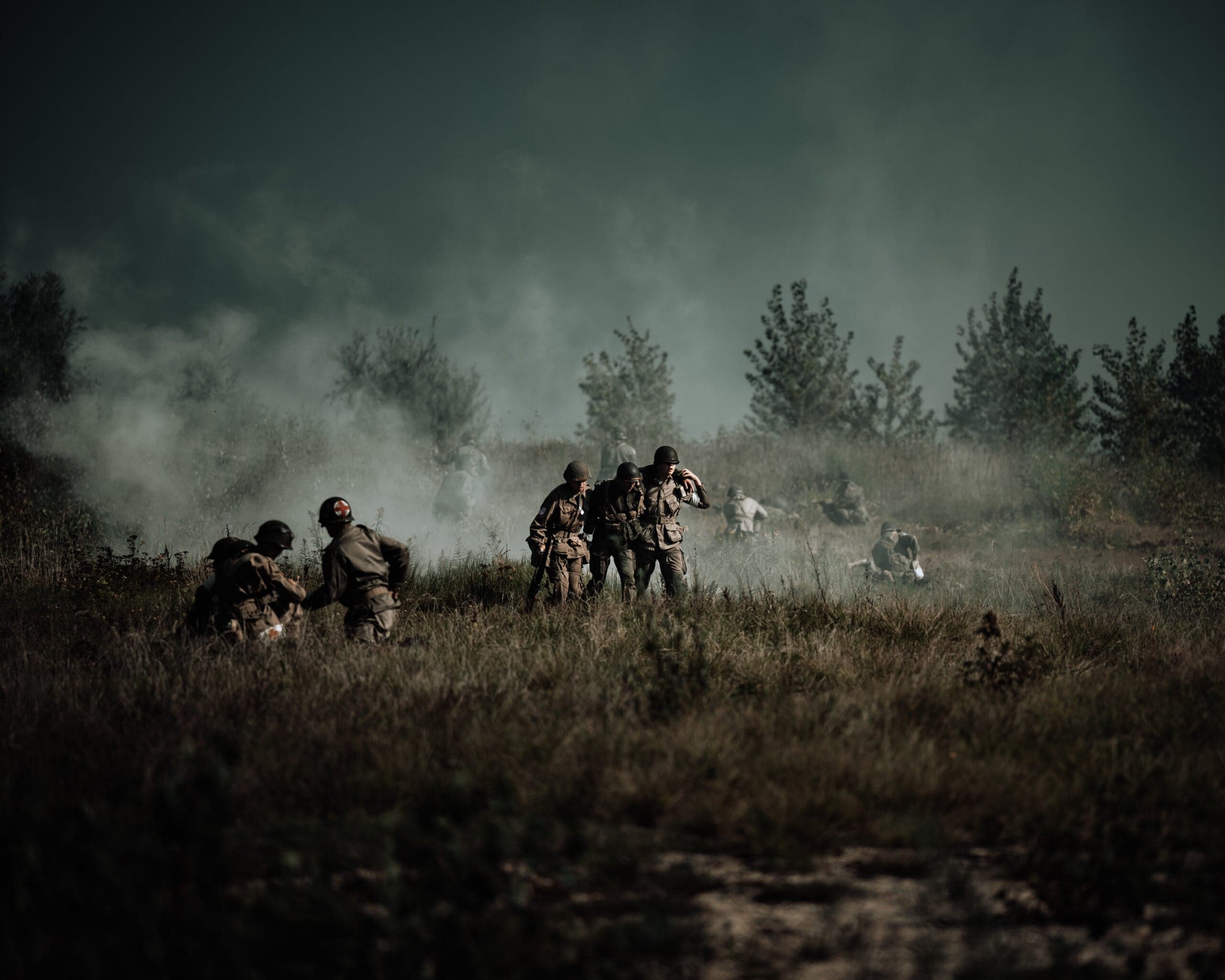 WO2-reenactment op een mistig veld: medic met rode-kruis-armband ondersteunt een gewonde kameraad, rook drijft door een grijsgroen landschap