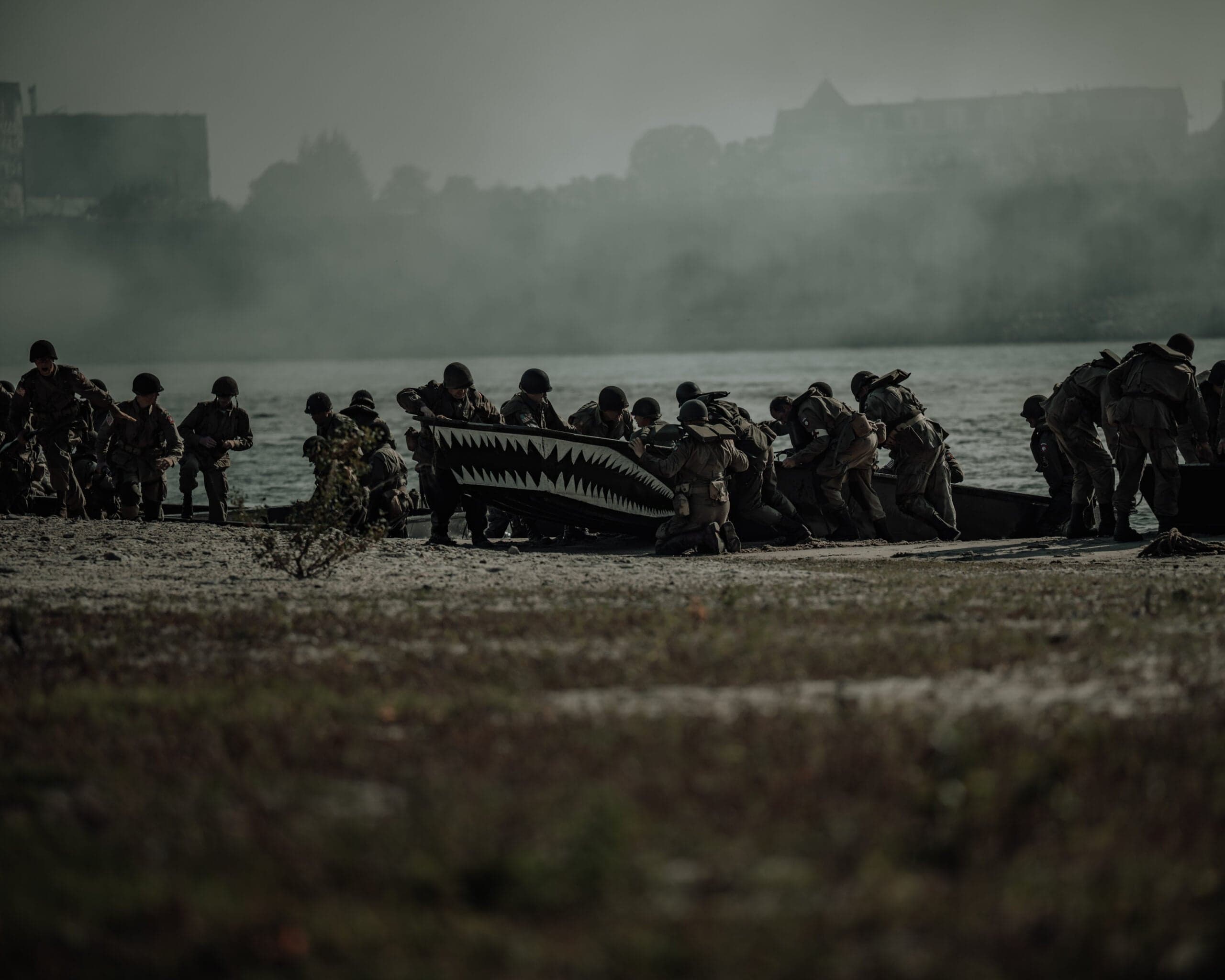 Groep soldaten sleept een landingsboot met geschilderde haaibek door het ondiepe water, dichte mist over de rivier op de achtergrond