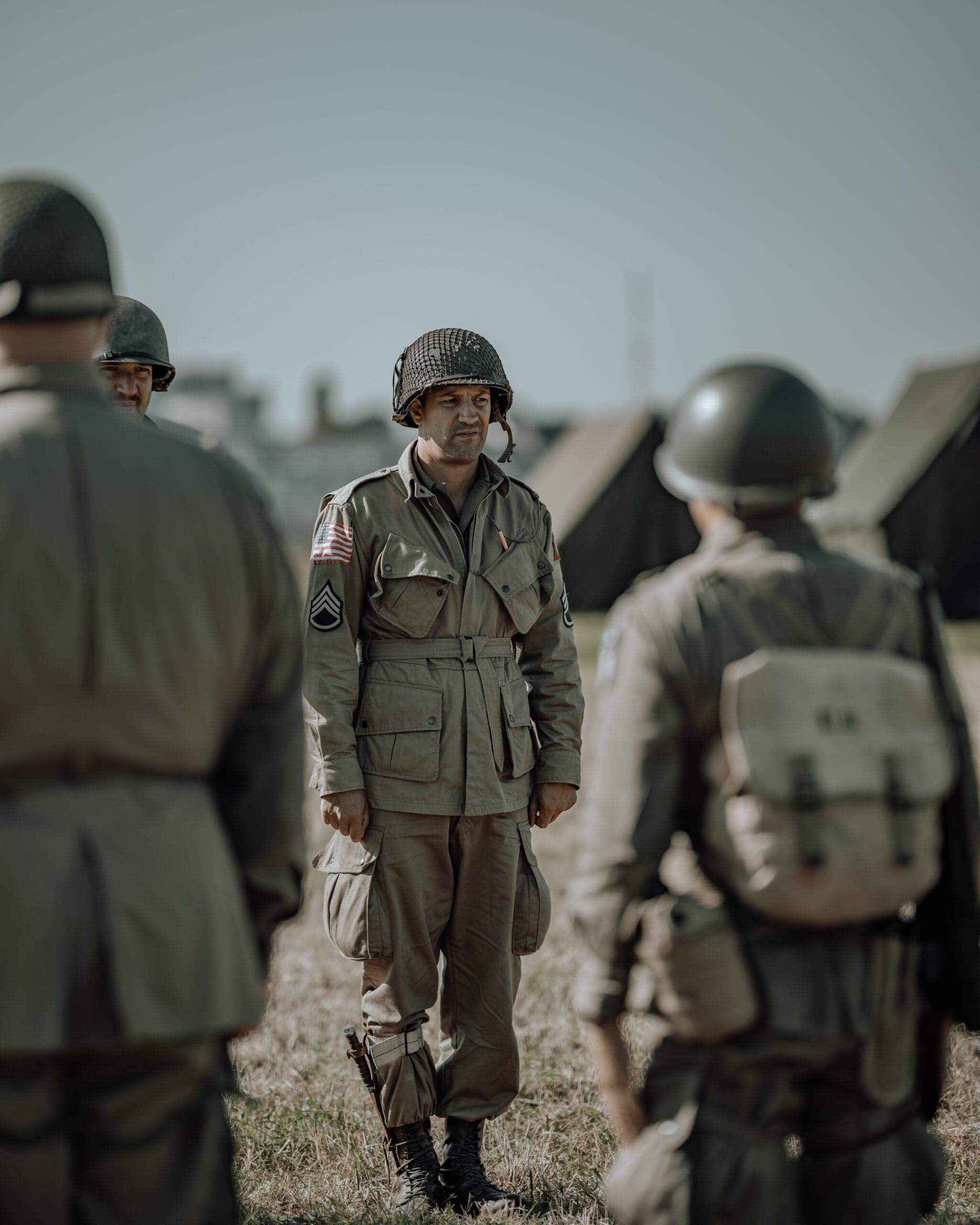 Amerikaanse paratrooper in volledig veldtenue met helmnetting, tussen twee collega's in een tentenkamp, alert zijdelings kijkend