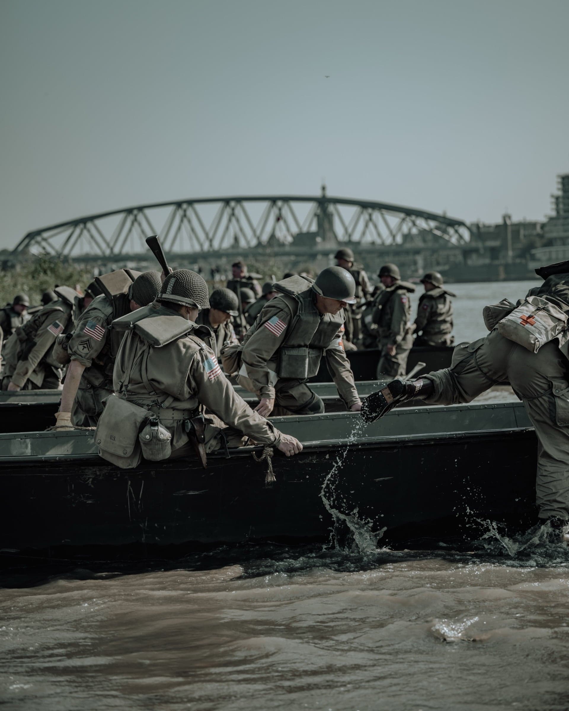 Dichte rij soldaten peddelend in een assaultboot op de Waal, de bogen van de Waalbrug en silhouet van Nijmegen op de achtergrond