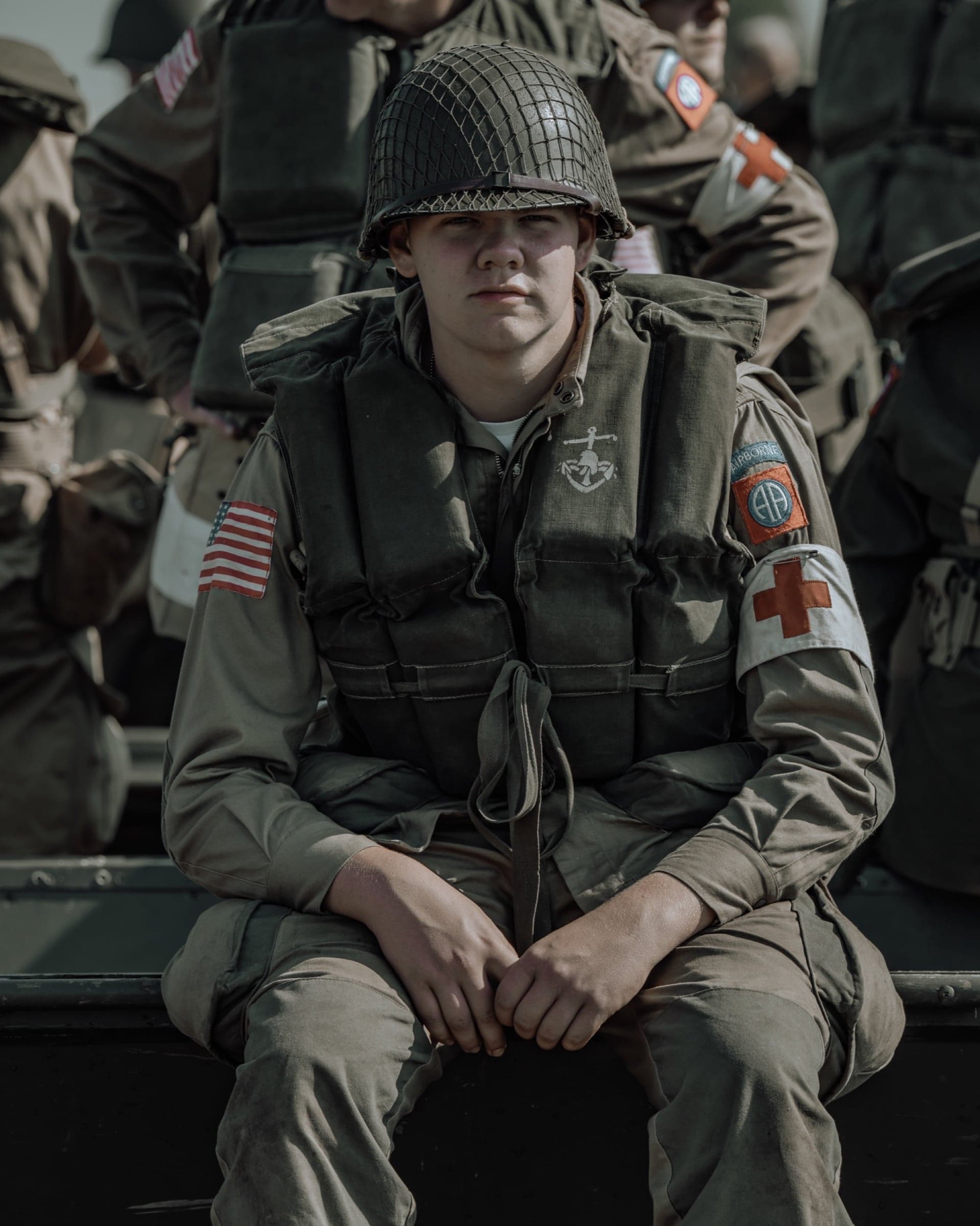 Zittend portret van een jonge medic met zwemvest en rode-kruis-armband, directe ernstige blik, andere soldaten onscherp op de achtergrond