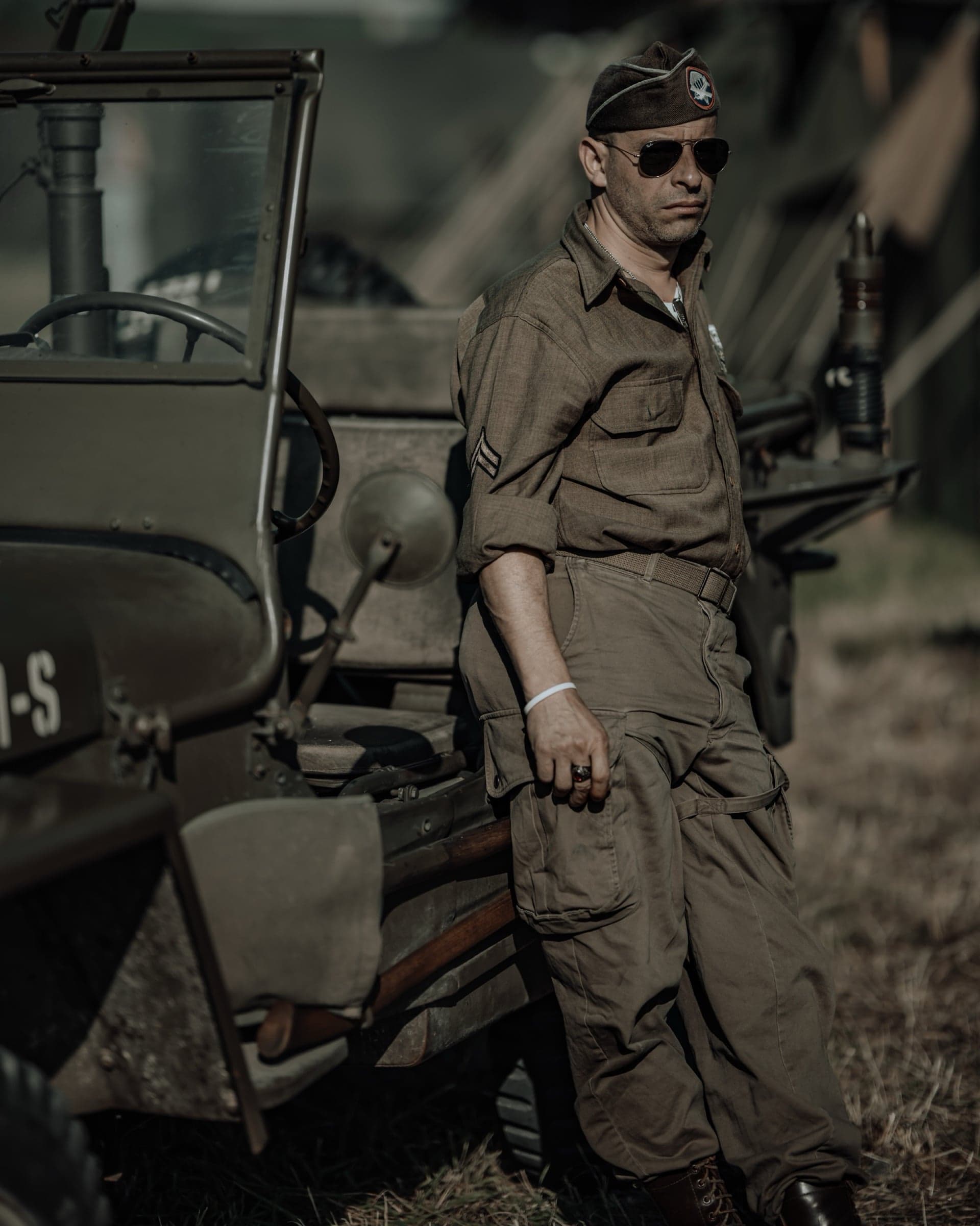 Amerikaanse airman in overseas cap en spiegelbril leunend tegen een Willys Jeep, tentenkamp in zacht bokeh op de achtergrond