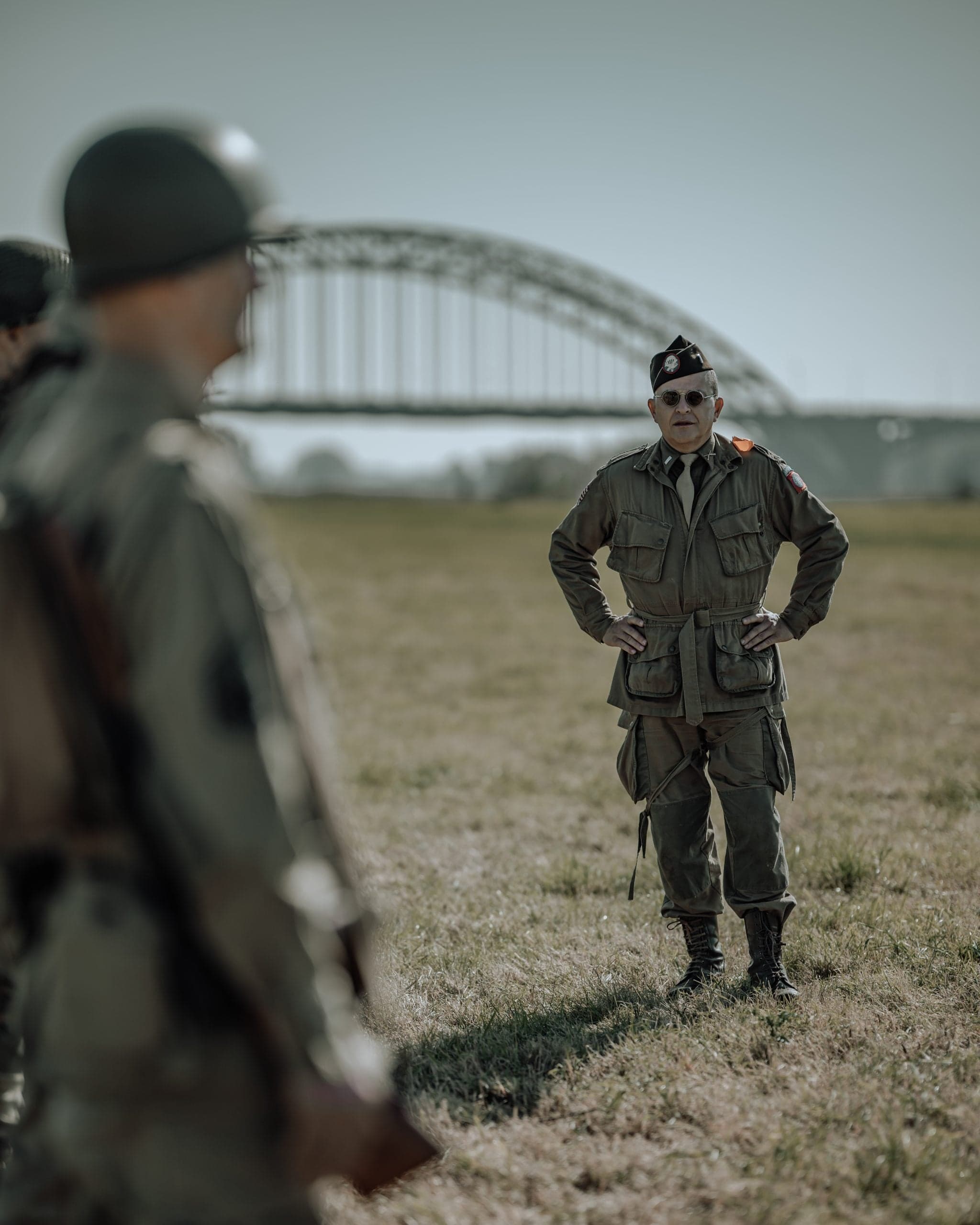 Amerikaanse commandant met handen in de zij onder de boog van de Waalbrug, twee soldaten op de voorgrond onscherp luisterend