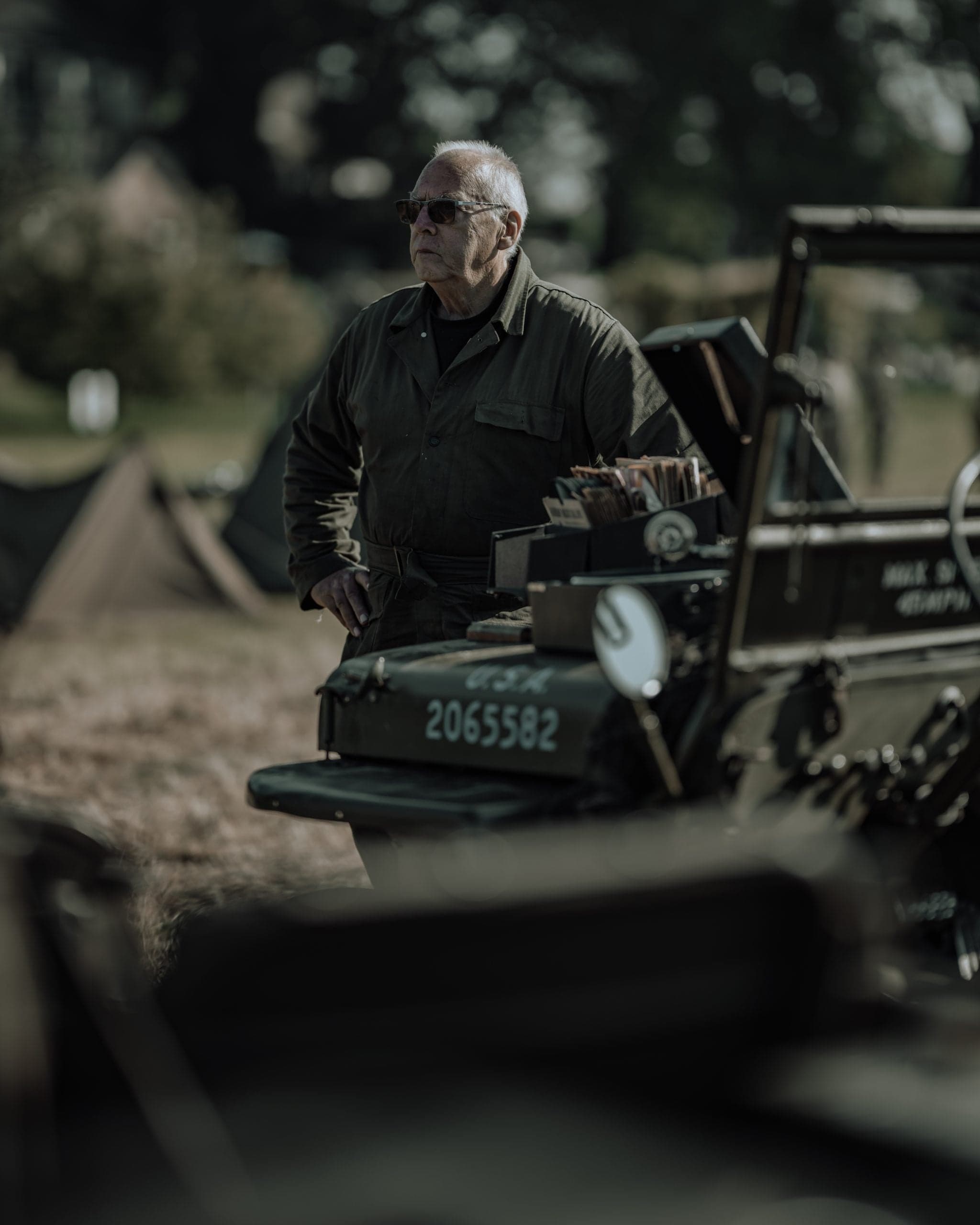 Oudere veteraan met spiegelbril en donkere coverall naast een US Army Jeep met nummer 2065582, stoffig middaglicht en tenten op de achtergrond