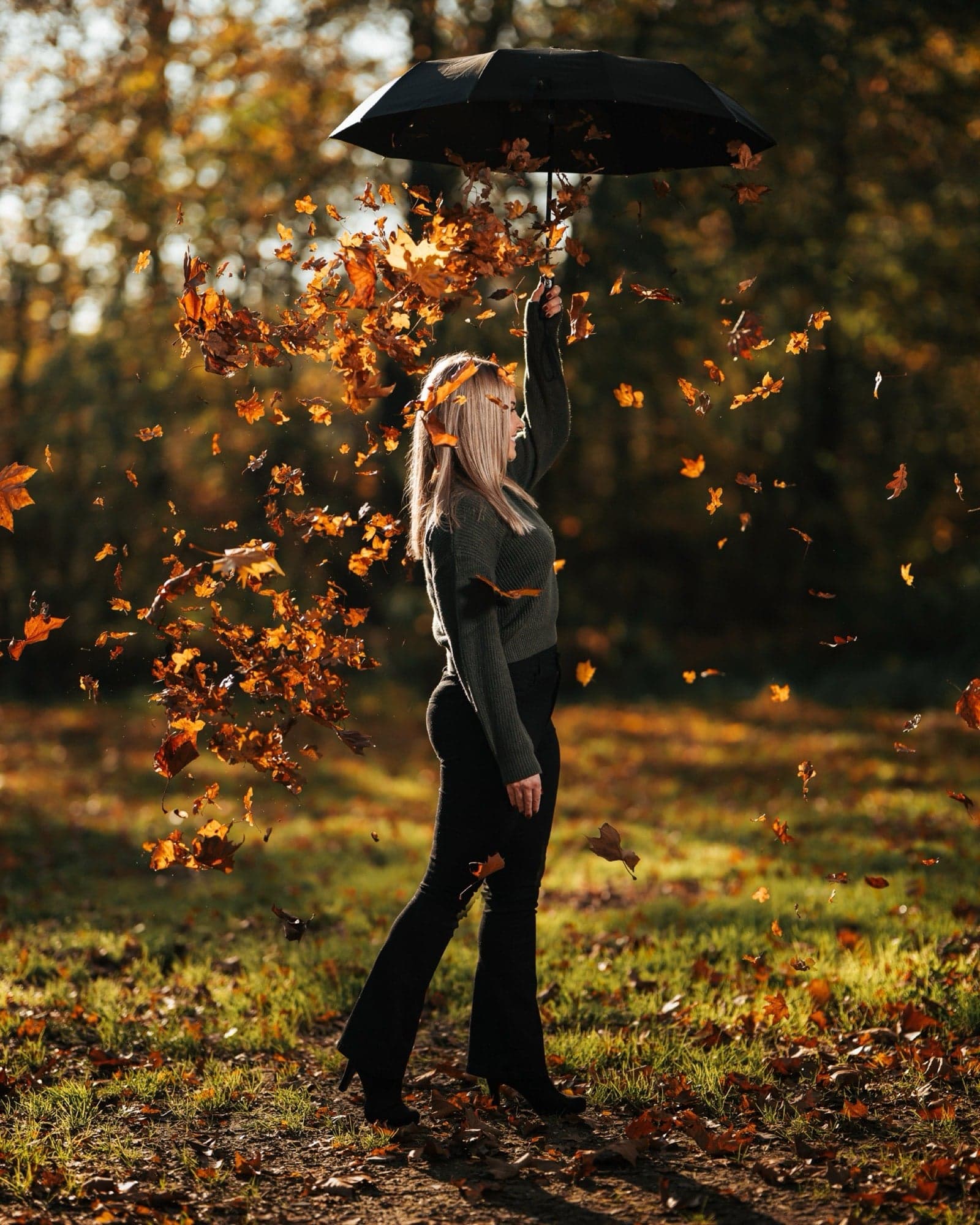 Vrouw danst met open paraplu door dwarrelende herfstbladeren in warm tegenlicht, silhouet tegen een herfstbos met gouden bokeh