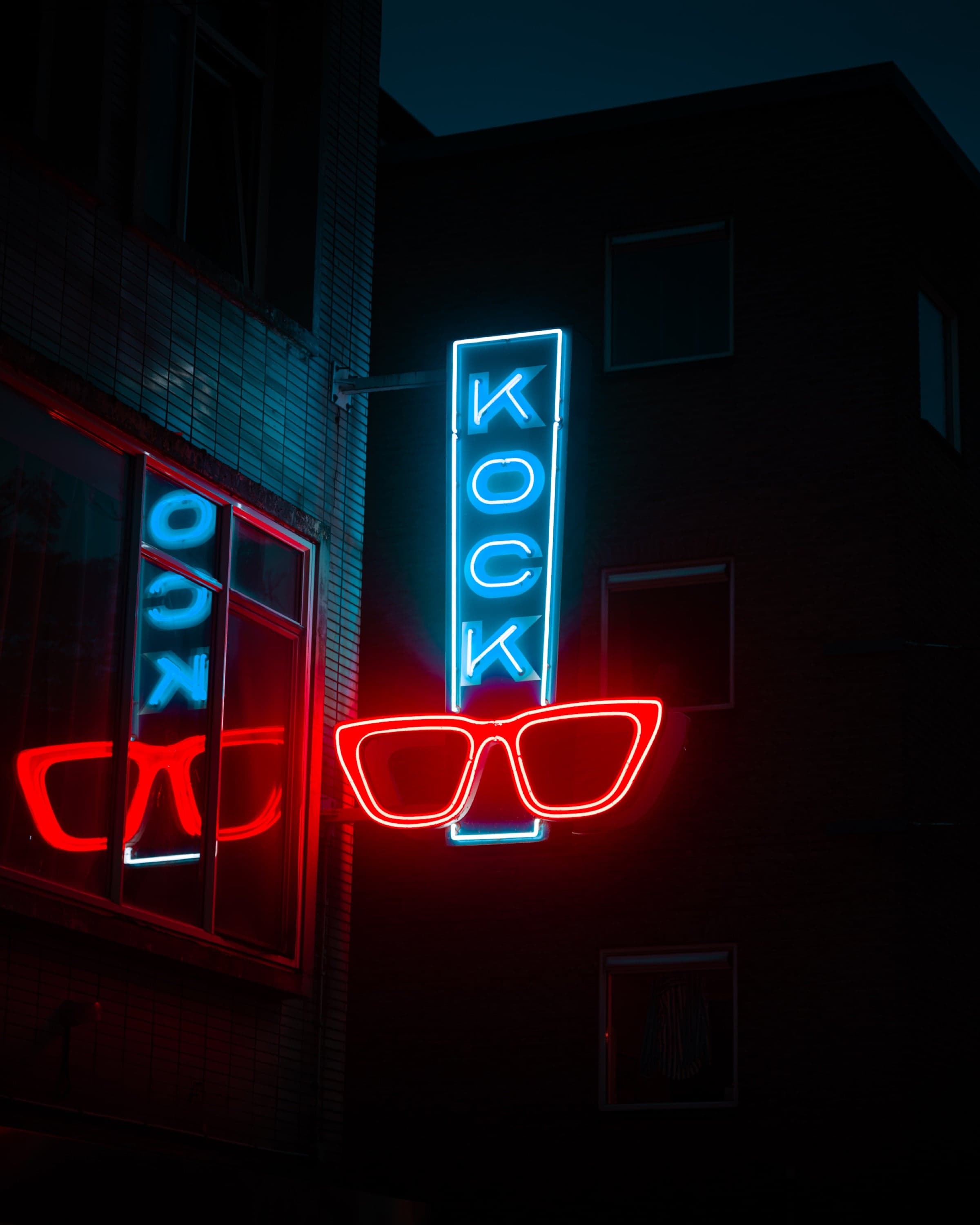 Neonreclame van een opticien 's avonds: blauwe letters KOCK boven een rode brilmontuur, gespiegeld in een naastgelegen etalage