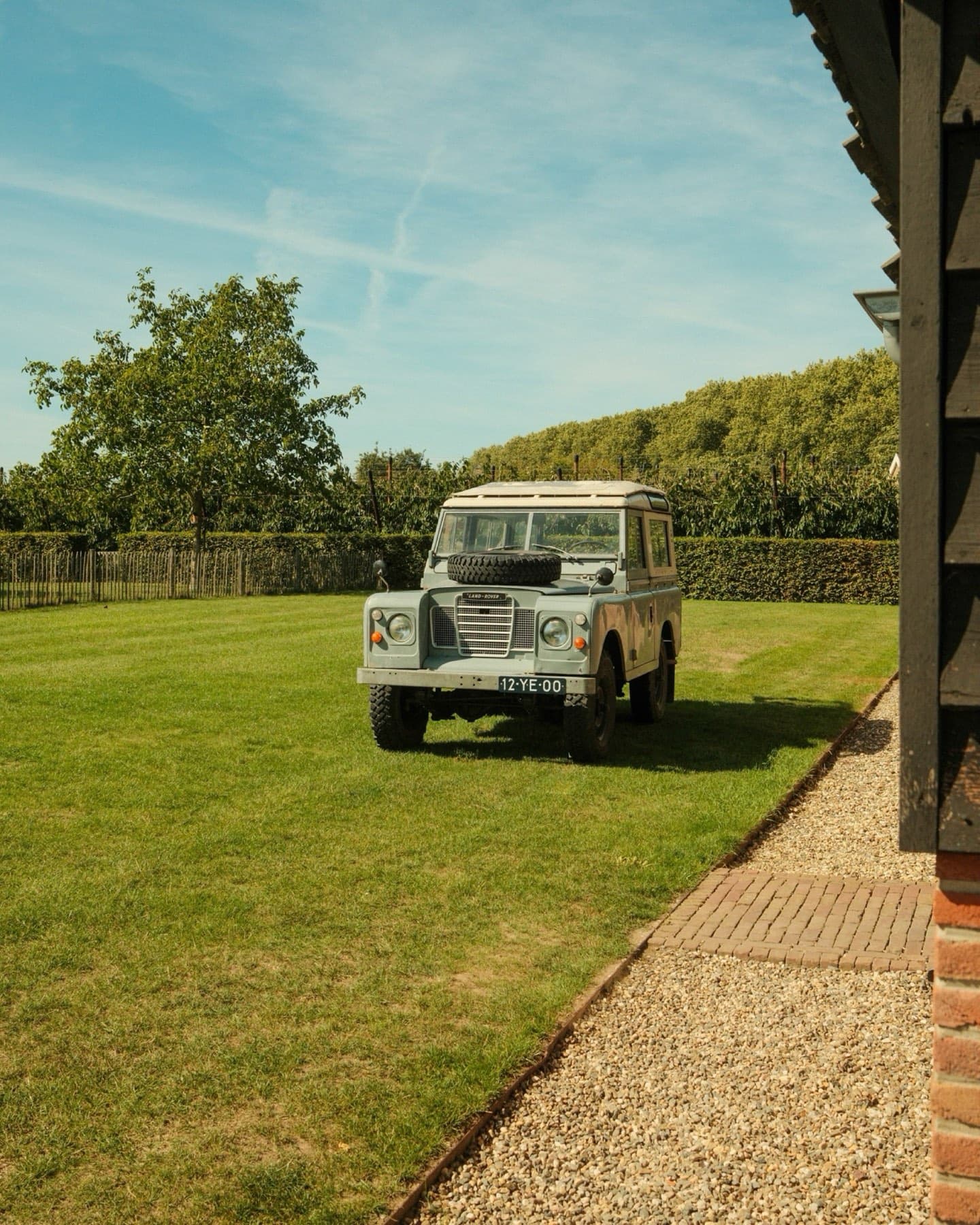Klassieke lichtblauwe Land Rover Defender geparkeerd op een frisgroen gazon naast een strakke hedge, onder een heldere blauwe zomerlucht